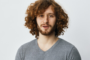 Young man with curly hair expressing confidence in a casual gray sweater, standing against a...