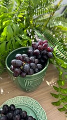 A close-up of fresh, dark purple grapes with water droplets, arranged in a green ceramic cup and plate. Vibrant green leaves add contrast, with a woven basket in the softly blurred background.