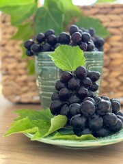 A close-up of fresh, dark purple grapes with water droplets, arranged in a green ceramic cup and plate. Vibrant green leaves add contrast, with a woven basket in the softly blurred background.