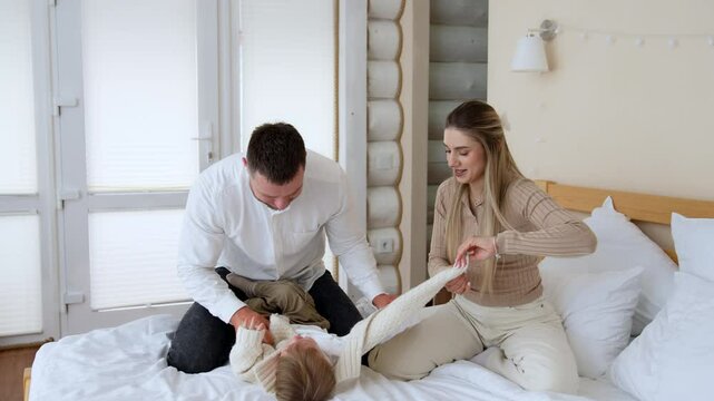Father tumbles his baby boy putting him on the bed. Parents take off kid's warm sweater. Beautiful family portrait.