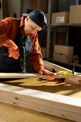 Female Carpenter drilling hole into timber block working in her workshop. Woman doing furniture assembling in studio. Woodworking expert using electric power drill tool.