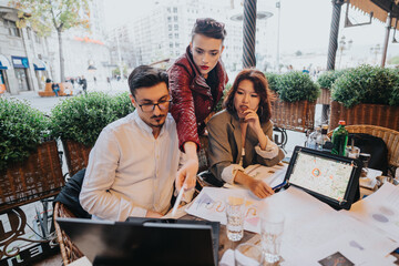 A diverse group of business people collaborating over laptops and documents at an outdoor cafe, generating ideas for a project. The setting fosters innovation and teamwork in a casual environment.