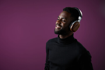 Young Black man enjoying music with headphones, smiling against a purple background, representing...