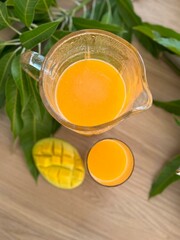 A fresh mango feast: a green plate holds diced and sliced ripe mango, next to a glass and pitcher of vibrant orange mango juice. Green mango leaves add a natural touch to the wooden table setting.
