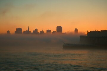 Fog blankets a city skyline at sunrise with silhouettes of buildings against a colorful sky