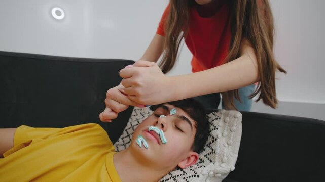 A girl smears toothpaste on her sleeping friend's face, filming the prank for social media. In colorful casual clothes, she captures the mischievous moment in a bright apartment