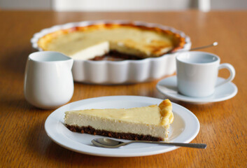 Piece of homemade cheesecake on a white plate on wooden table, a cup of black coffee with saucer, a cup of milk and a baking dish in the background. Coffee nad cake, side view, selective focus.