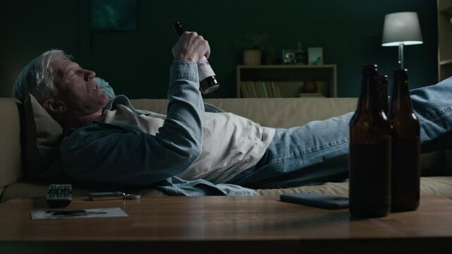 Low key shot of drunk elderly Caucasian man lying on couch in living room in front of empty alcohol bottles, finishing another beer and dropping bottle on messy floor spending evening alone
