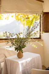 A minimalist arrangement of olive branches in a vase placed on a white-draped table near a window, with golden sunlight streaming in. A serene and elegant decor piece