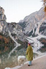 A serene moment of a woman with her dog by the calm lake in the Dolomites. The dramatic mountain reflections on the water and the autumn landscape create a breathtaking natural scene