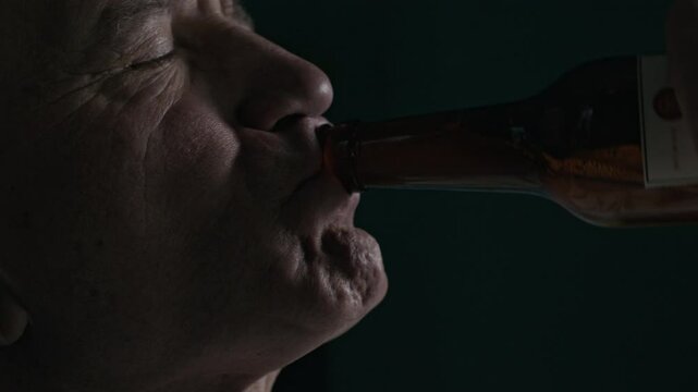 Closeup low key shot of gloomy mature Caucasian man with drunk eyes gulping down bottle of beer and looking off into distance with numb expression