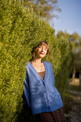 Portrait of a woman leaning against a tall, lush hedge, enjoying the sunlight. She is dressed in a...