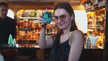 Beautiful woman guest zipping to taste in Christmas blue Lagoon cocktail with good mood sitting on luxurious counter bar green neon snow Christmas pine tree festive theme on wine background. Vinosity.