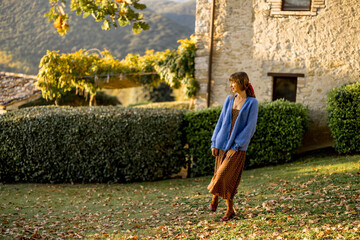 A stylish woman walking on a grassy lawn near a rustic stone house, surrounded by autumn colors and...