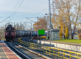 A red locomotive pulls a freight train on curved tracks at a station with a platform, yellow railings