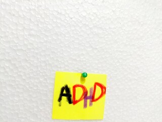 A yellow paper note with the abbreviation ADHD on it pinned to a cork board. Close up. ADHD is...