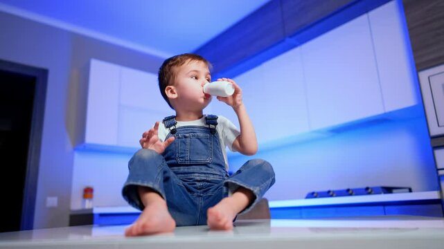 Adorable male kid in jeans romper sits on the table snacking. Cute boy drink yogurt from a bottle. Low angle view.