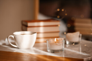Burning candle with cup of coffee and stack of book on marble table in room. Cozy home atmosphere. Good morning.