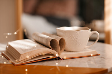 Cup of tea staying on open paper book with folded pages in heart shape on marble table with glowing lights close up. Valentines Day.