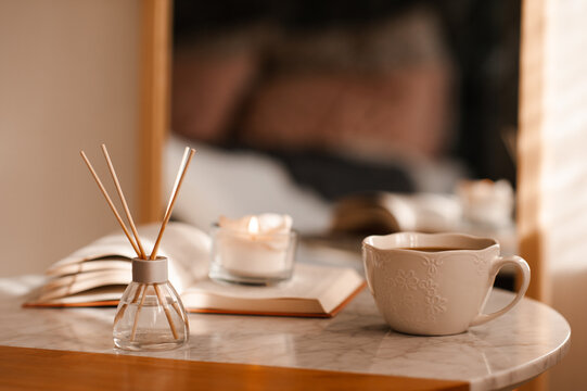 Cup of coffee with aroma diffuser and burning candle on paper book staying on marble table in room close up. Cozy home atmosphere.