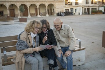 Senior friends reading a map together on a bench in a picturesque Spanish plaza during a cultural...