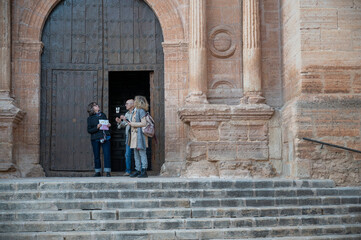 Three senior tourists standing on historic stone steps, discussing a map in front of a grand wooden church door, in a spanish village