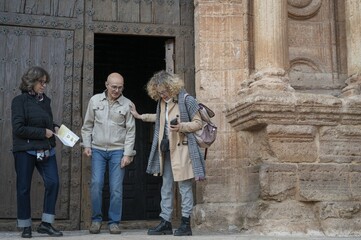 Three senior friends exiting a historic Spanish church while holding a map and engaging in a cultural sightseeing tour