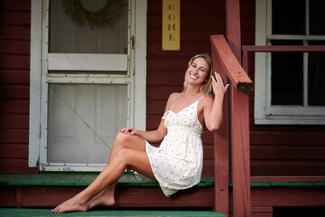 Sexy blonde woman sits in short white sundress on front porch of house at dusk