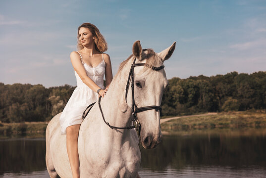 Young woman in white dress riding a horse by a lake in nature