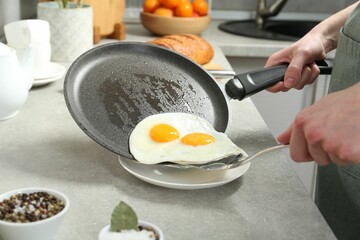 Woman putting tasty fried eggs onto plate from frying pan in kitchen, closeup