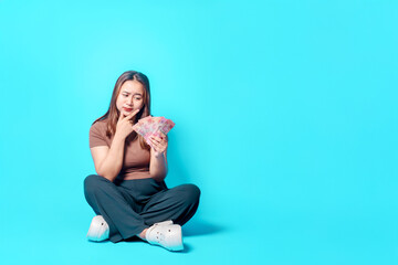 A woman holds cash while sitting cross-legged against a vivid blue background contemplating finances