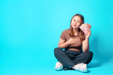 A joyful young woman sits cross-legged while clutching banknotes close to her with a delighted...