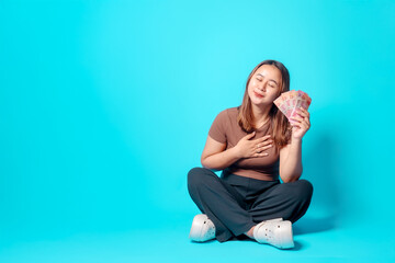 A joyful young woman sits cross-legged while clutching banknotes close to her with a delighted...