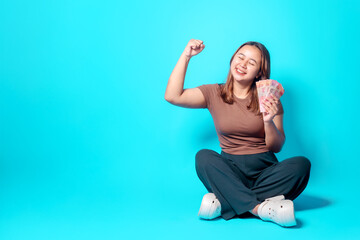 Fototapeta premium A cheerful young woman sits on a blue background, excitedly holding a fan of cash and raising her fist in a victorious gesture, symbolizing financial success, achievement, and positive emotions.