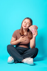 A joyful young woman sits cross-legged while clutching banknotes close to her with a delighted...