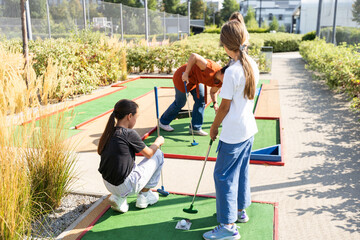 Cute school girl playing mini golf with family. Happy toddler child having fun with outdoor activity. Summer sport for children and adults, outdoors. Family vacations or resort.