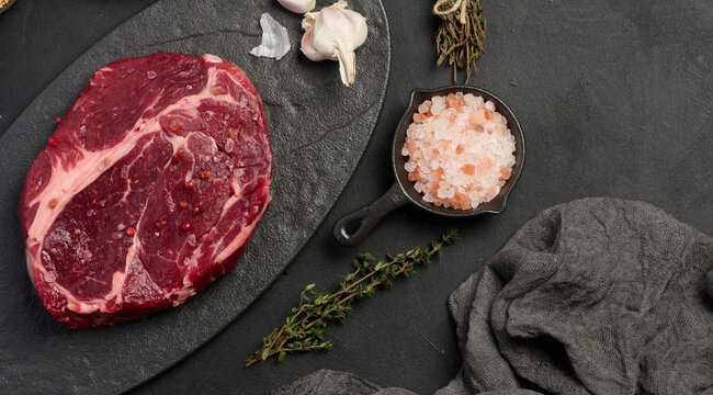 Raw beef steak chuck eye roll on a blck board. Spices for cooking Himalayan salt, peppercorns and thyme sprigs