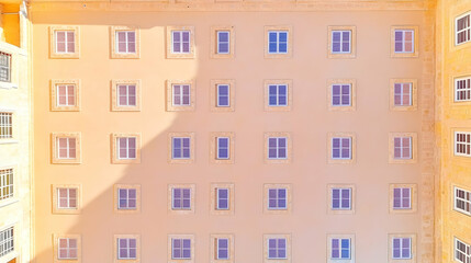 Naklejka premium Symmetrical building facade with numerous windows, viewed from above, showcasing a pattern of light and shadow on a pale yellow wall.
