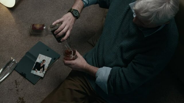 Top view low key shot of drunk senior Caucasian man sitting on floor with empty bottles and photo of ex-w, pouring last drops of whiskey suffering from painful breakup in messy home