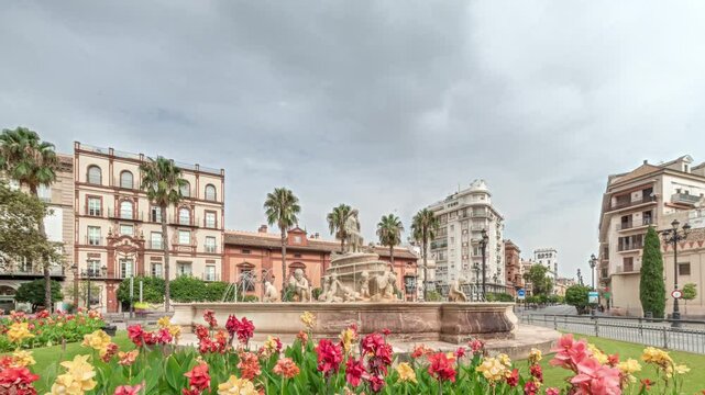 Hispalis Fountain in Seville, one of the city's most beautiful fountains, surrounded by colorful flowers and historical buildings. Timelapse hyperlapse with a cloudy sky in the background.