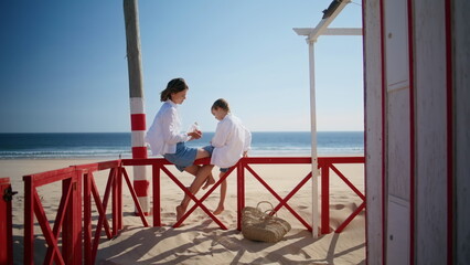 Happy woman talking son sitting beach red fence sunny day. Relaxed mother child