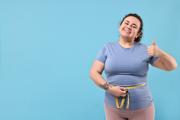 Weight loss. Happy plus size woman measuring waist with tape and showing thumbs up on light blue...
