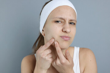 Upset teenage girl popping pimple on her face against grey background. Acne problem