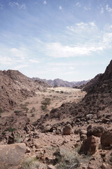 A vast Saudi desert valley surrounded by rugged mountains under a bright blue sky. The rocky landscape stretches endlessly, revealing a peaceful, untouched part of the Arabian wilderness.