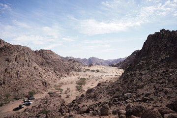 A vast Saudi desert valley surrounded by rugged mountains under a bright blue sky. The rocky landscape stretches endlessly, revealing a peaceful, untouched part of the Arabian wilderness.