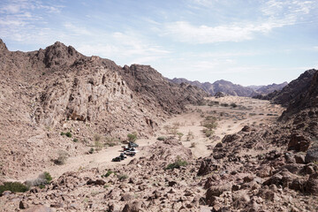 A wide view of rugged Saudi desert mountains surrounding a dry valley with scattered trees and parked off-road vehicles. A dramatic natural landscape showcasing the raw beauty of the Arabian wildernes