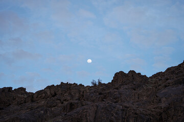 The moon rises above rugged Saudi mountains under a soft blue sky scattered with light clouds. A peaceful desert landscape capturing the natural beauty and calm atmosphere of the Arabian wilderness.