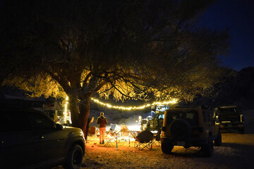 A cozy desert campsite at night, illuminated by string lights wrapped around a tree. Off-road vehicles and camping chairs surround a warm fire, creating a peaceful and adventurous outdoor atmosphere.