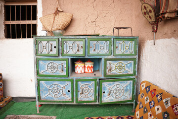 Traditional Saudi interior featuring a hand-painted wooden cabinet with geometric motifs, set against a mud-brick wall and surrounded by woven cushions, reflecting authentic Saudi heritage and decor.