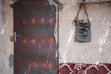 Traditional painted door with colorful star motifs beside a textured beige wall and an old leather bag hanging above patterned seating. A rustic cultural scene reflecting heritage and handcrafted desi